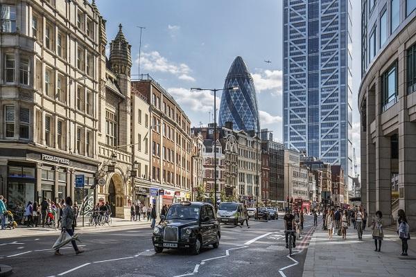 A shot of the financial district in the City of London, with a black cab, the Gherkin and people walking along the footpath.