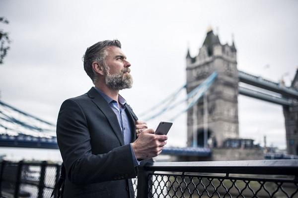 A business man with a grey beard talking on the phone while leaning on a railing next to the Thames in London.