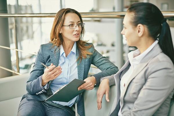A woman being coached on her career in an open plan office by another woman wearing glasses and holding a clipboard.