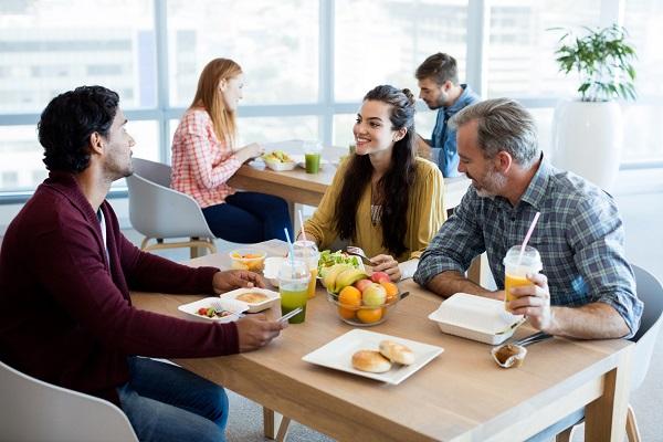 A shot of three colleagues around a table in an open plan office eating a free lunch that has boosted their employee morale.