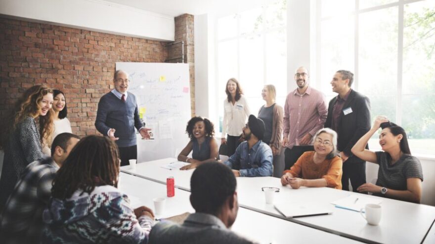 A group of colleagues in a business meeting, most laughing and smiling while discussing company matters.