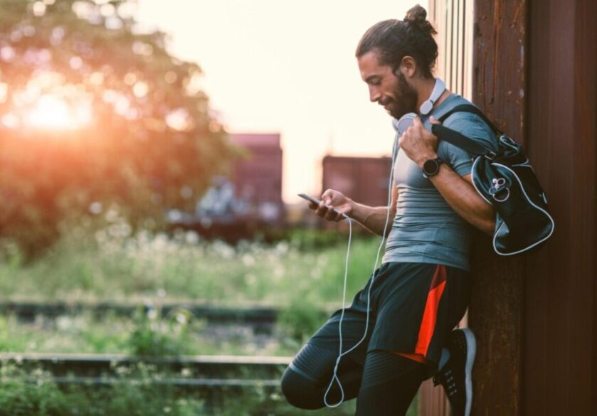 A man looking to recruit a virtual staff member with his phone, leans against a wall outside while wearing exercise gear.