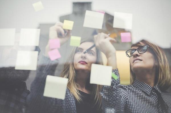 Two business women in fashionable clothes using and writing on post-it notes on a glass wall while in a meeting.