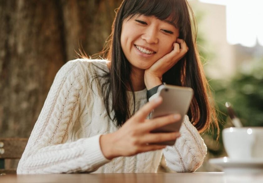 Happy young woman sitting at an outdoor cafe looking to recruit and using her cellphone.