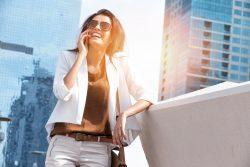 Woman in a white jacket and sunglasses talking on phone nearby Dubai’s Jumeirah Lake Towers.