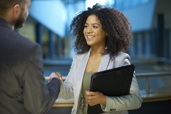 A hiring manager is shaking hands with a colleague while standing in an office building and holding papers.