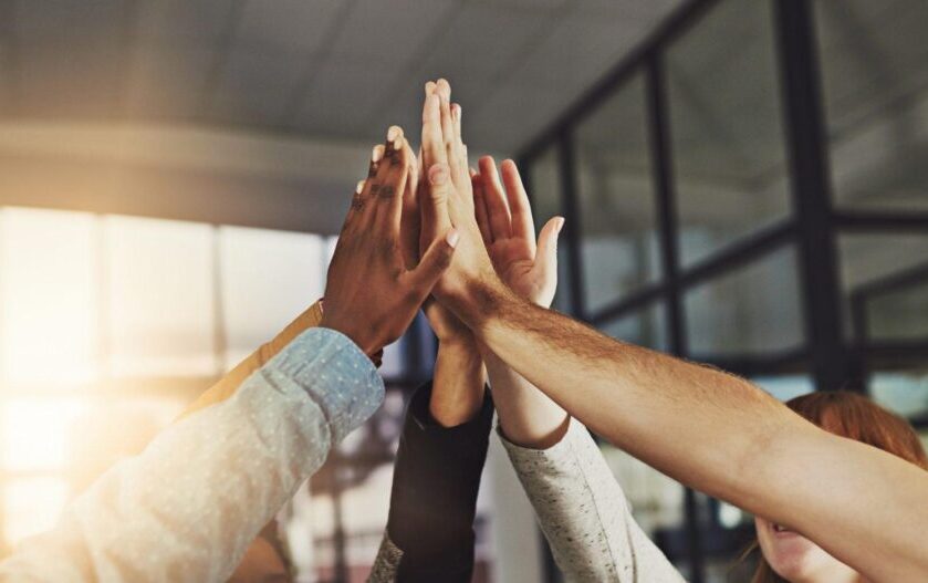 Shot of a group of five colleagues giving each other a high five in a well-lit office with windows in the background.