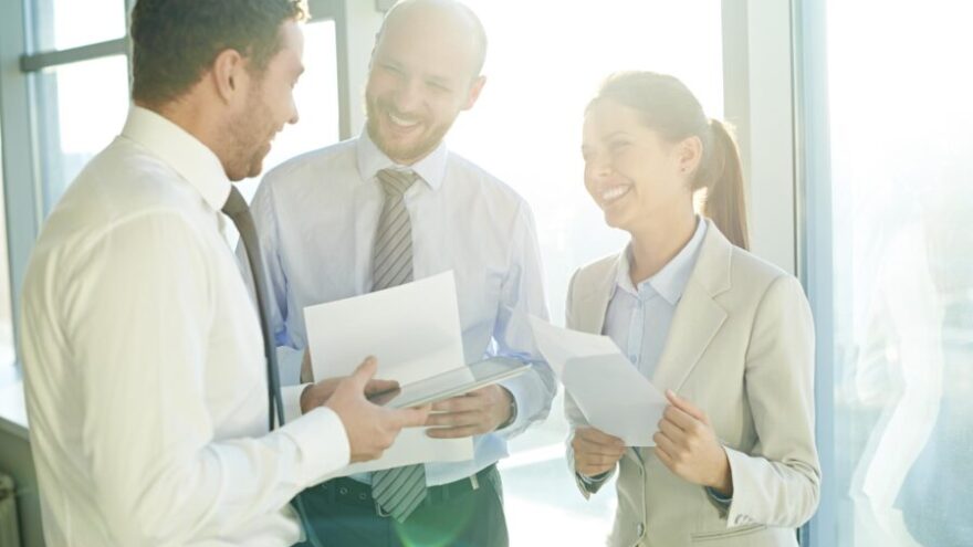 Three colleagues smiling together while in a meeting next to the windows of a well-lit office building.