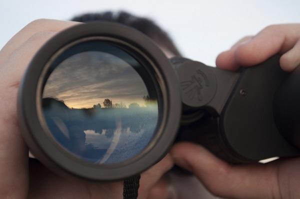 A close up on a man holding binoculars looking out on a lake at sunset.