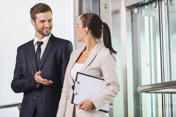 A man talking to his female colleague about his elevator pitch outside an elevator in their office.