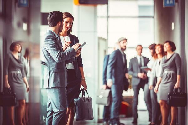 A recruiter talking to a job seeker while standing in an office. In the background, there are several business people.