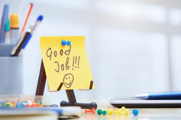 A post it note that says ‘good job’ with a smiley face on a desk with pens and thumb tacks spilt on the surface.