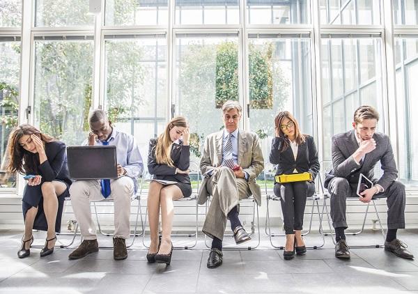 Six people sitting along a window in a building waiting to interview for their ideal job.