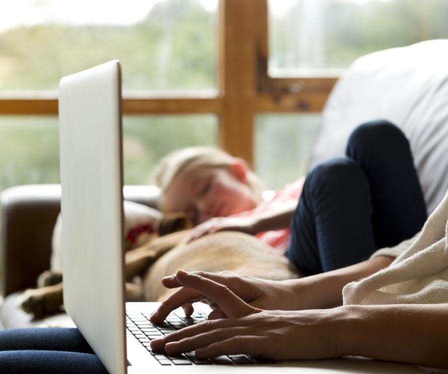 Home worker works on her laptop on the sofa while her child sleeps in the background