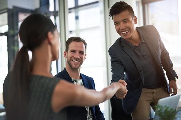 A male candidate for a PA job interview smiles and shakes hands with the interviewer in a sunlit office.