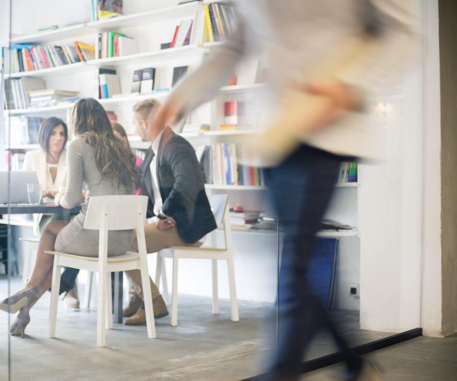 Group of creative people on a meeting in office, blurred motion of businessman passing in front, view through window