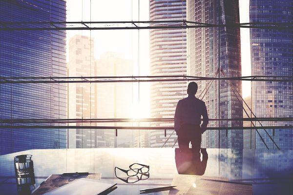 A man looking out into a city skyline from a glass-lined office, thinking about quitting his job.