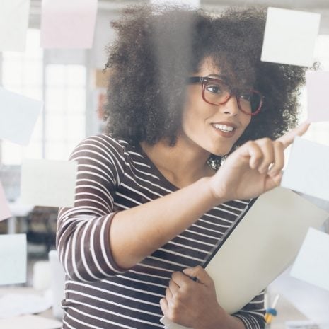 A private PA with glasses puts post-it notes on a window to organise her tasks while holding a bundle of papers.