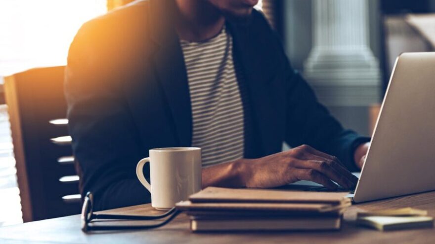 Close-up part of VA is using a laptop while sitting at his working place at home, with glasses and a coffee mug next to him.