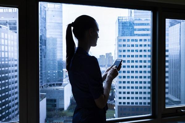 A woman uses a mobile phone to search for jobs while standing near a window, looking out at a city skyline.
