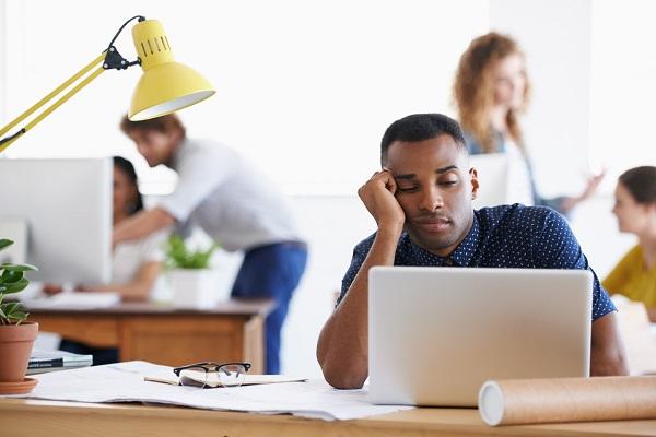 A sad worker sitting in an open plan office in front of his laptop while his colleagues work in the distance.