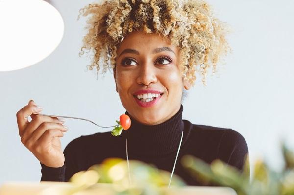 Woman eating a cherry tomato and lettuce to maintain a healthy office lifestyle while smiling and looking away.