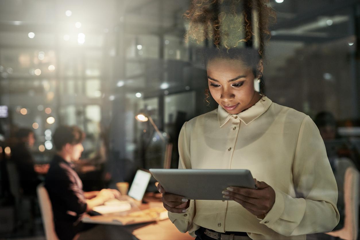 A general manager looks at her ipad while workers sit at their desks in the background