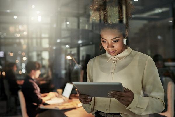 A private PA using an iPad in the foreground while her colleagues work in the background of an office at night.