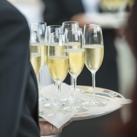A temporary waiter carrying seven champagne glasses on a tray at a party.