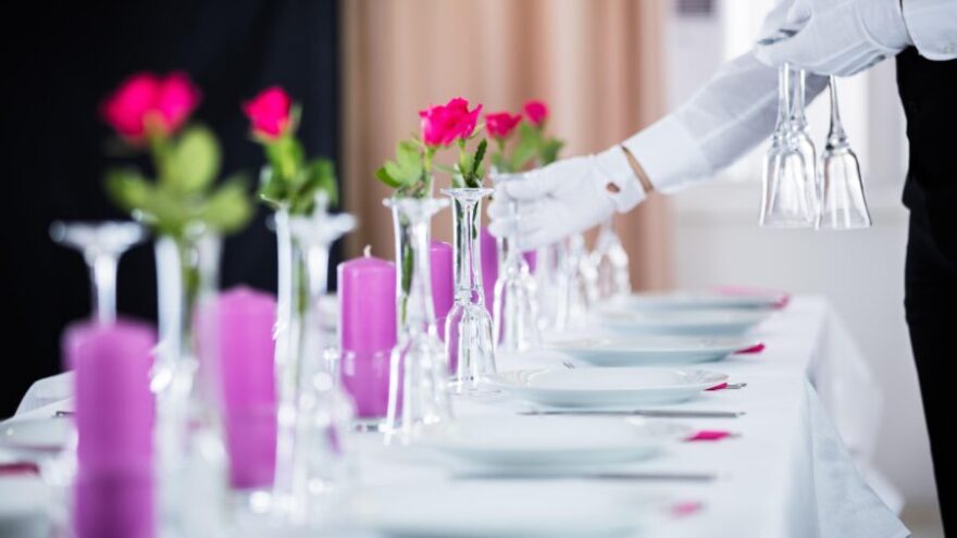 A butler places water glasses on a white tablecloth in a mansion, which is one of the qualities every butler should possess.