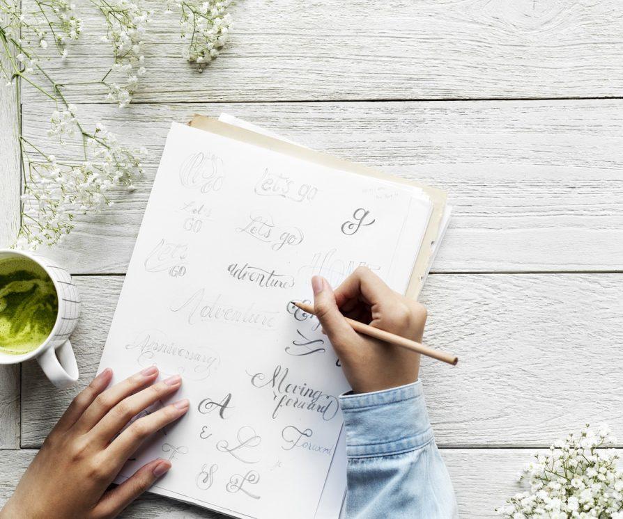 A woman's hands writing calligraphy in different styles while sitting outside on a picnic bench.