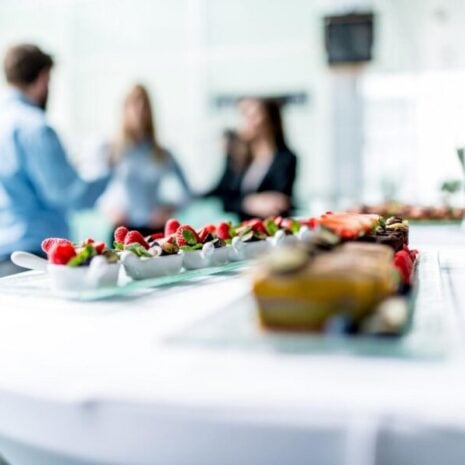 Closeup of two lines of canapés, with champagne glasses and three people in the background talking and socialising.