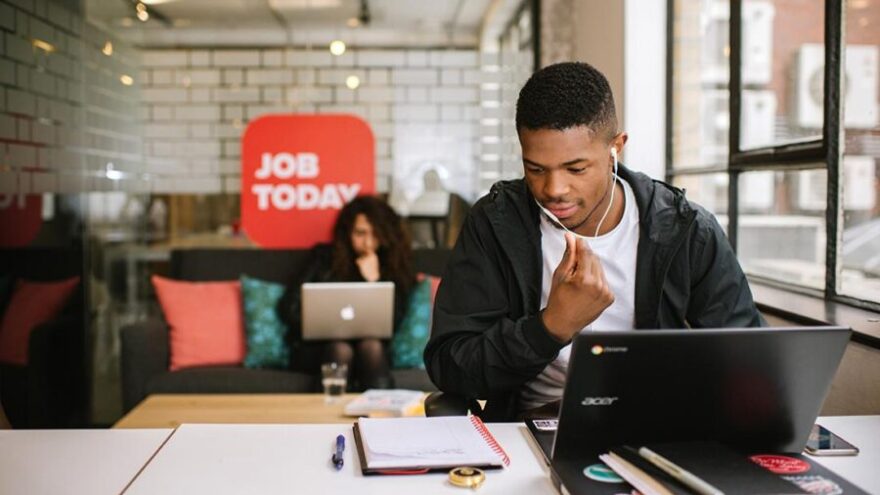 A man in an open plan office using a laptop and working near a window with a female colleague in the background.