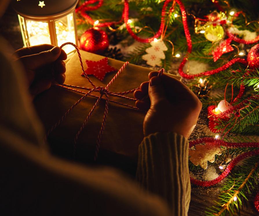Crop shot of person tying up colorful twine on wrapped Christmas present at table with glowing garland