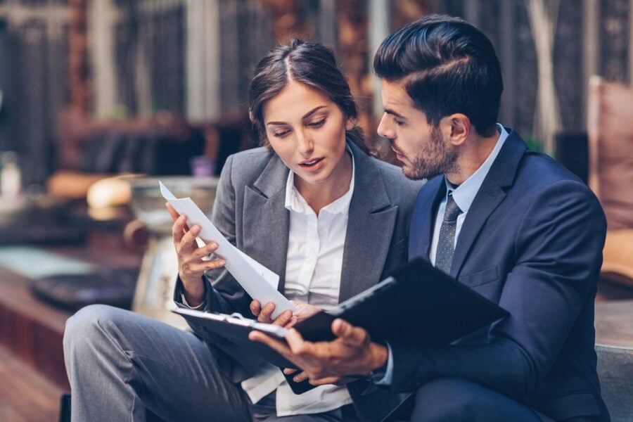 Two business people, one man and one woman, sitting on an outdoor step and talking about business documents.
