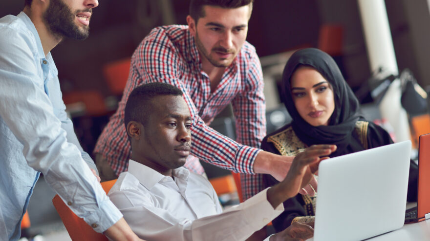 A group of four human resources staff working and talking in an office connected with tech devices like tablets and laptops.