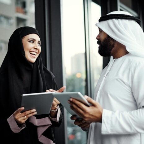 Two arab co-workers smiling and comparing notes outside an office in Dubai.