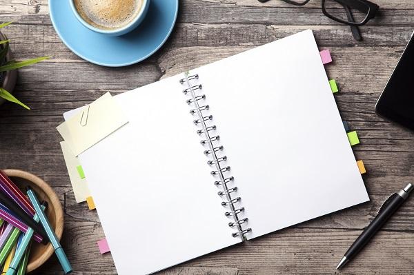 A notebook on a desk with a cup of coffee and a container of pens.