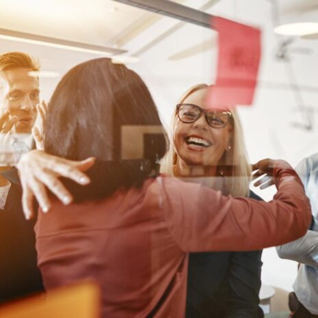 Two HR businesswomen are hugging and smiling while brainstorming with their colleagues in a bright, contemporary office space.