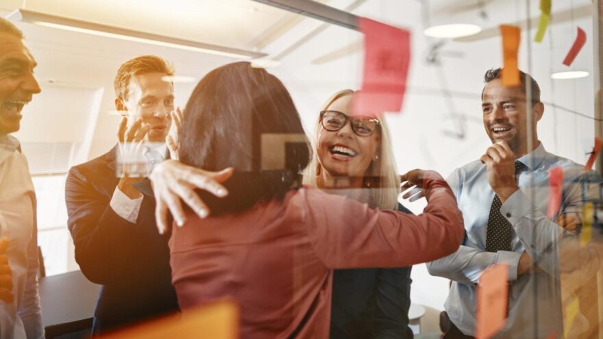 Two HR businesswomen are hugging and smiling while brainstorming with their colleagues in a bright, contemporary office space.