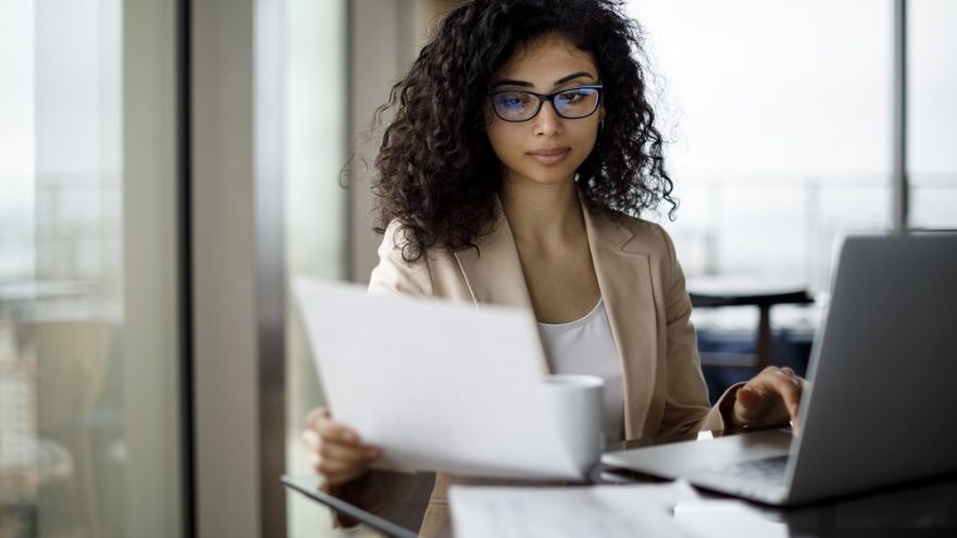 A young businesswoman working on a laptop while reading notes.