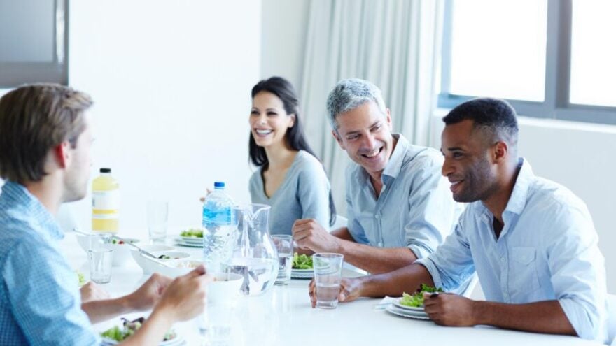 Four colleagues are also friends at work, laughing and talking around a table in a well-lit office.