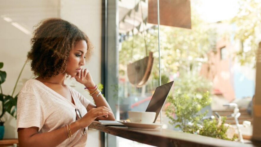 Young VA at a cafe using a laptop while looking outside onto the street in front of her. She’s also drinking a cup of coffee.