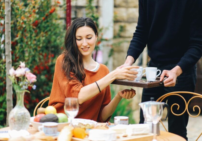 A young woman in an orange top sitting at a table outside being served by a modern-day butler.