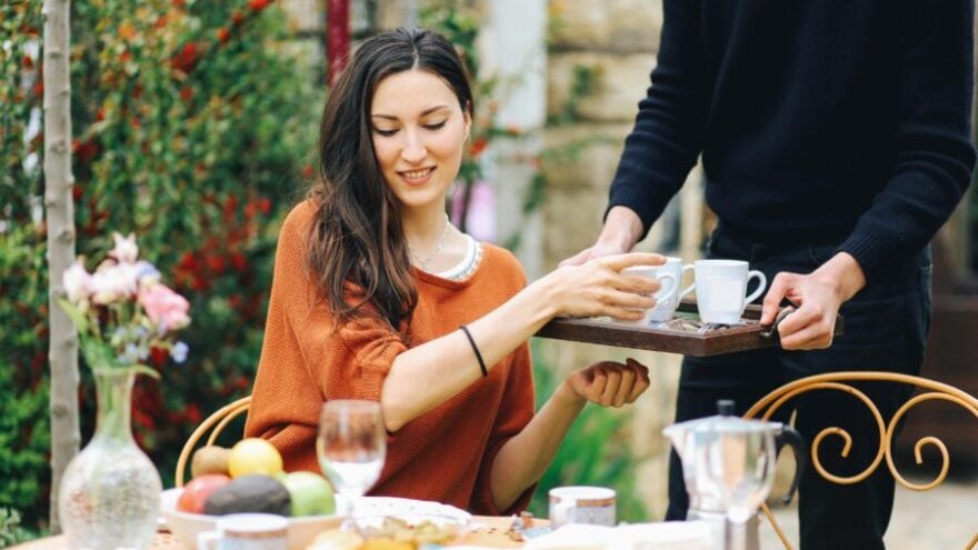 A young woman in an orange top sitting at a table outside being served by a modern-day butler.
