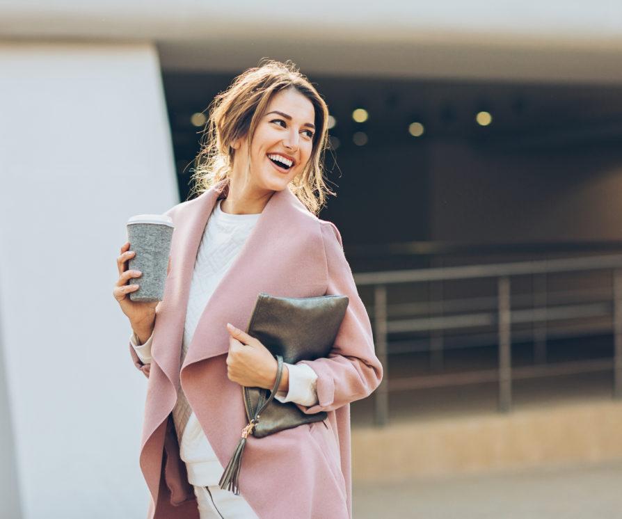 An elegant young temp wearing a pink coat walking on the sidewalk in an urban setting and looking back behind her shoulder.