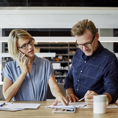 Two people working side by side, one on the phone and one looking at a report next to a coffee cup.