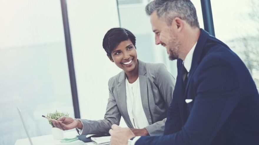 A business man and woman talking and smiling over a laptop computer while meeting in a modern office with windows for walls.