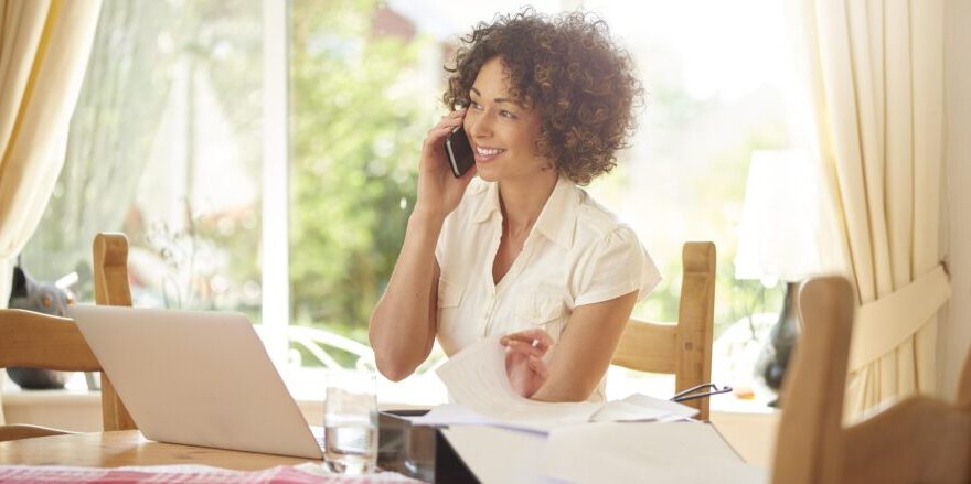 A self-employed woman gigging inside in her home office, talking on the phone and using her laptop.
