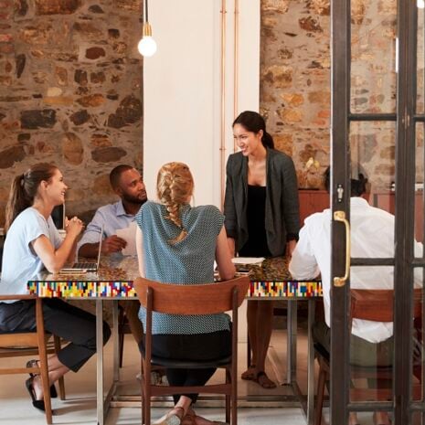 A human resources team of five people in a creative office with exposed brick wall and hanging lights having a group meeting